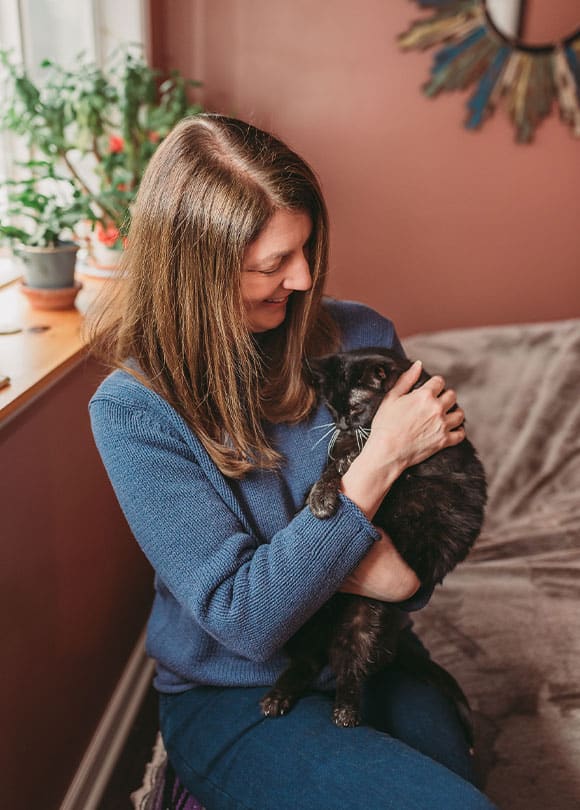 A woman with long brown hair, wearing a blue sweater, sits indoors by a window and smiles while cuddling a black cat like a caring vet. Plants and a decorative mirror are visible in the background.
