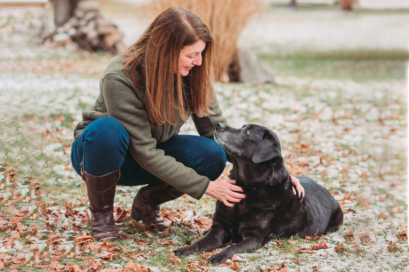 A woman with long brown hair crouches outside on autumn leaves, gently petting a black dog lying on the ground. The vet smiles as the dog looks up at her. There is light snow on the grass and trees in the background.