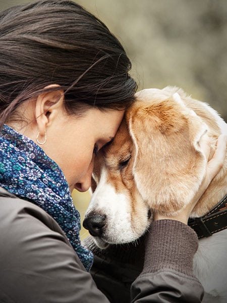 A woman, possibly a veterinarian, gently presses her forehead against a dog's head, both with eyes closed, sharing a tender, comforting moment. The woman holds the dog’s face in her hands as they appear calm and peaceful.