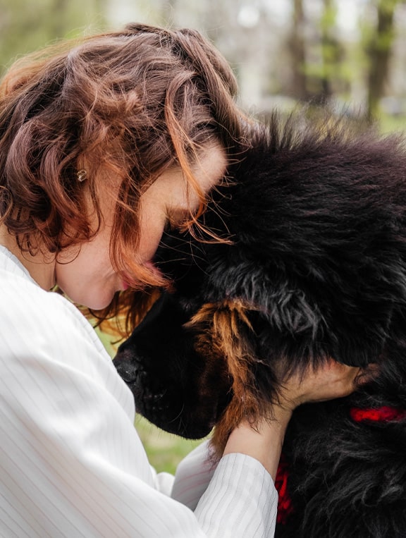 A woman in a white shirt, possibly a veterinarian, gently presses her forehead to a large, fluffy black and brown dog, sharing a tender, affectionate moment outdoors.