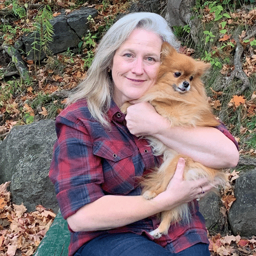 A woman with long gray hair in a red plaid shirt sits outdoors, smiling and hugging a small fluffy brown dog. They are surrounded by rocks and fallen autumn leaves.
