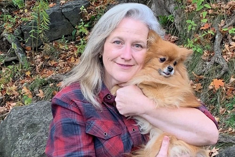A woman with long gray hair wearing a red plaid shirt smiles while hugging a fluffy brown Pomeranian dog outdoors with rocks and autumn leaves in the background.